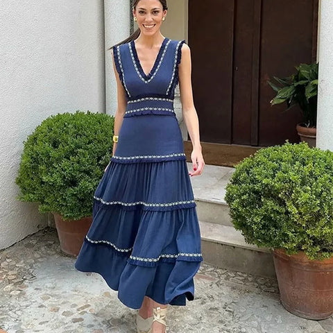 Woman wearing a blue dress with white trim standing outdoors next to potted plants.