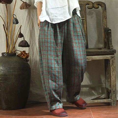 Person wearing plaid pants and a white shirt sitting on an old wooden chair with a vase and dried plants in the background.